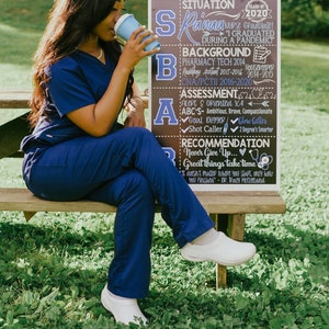 K&ouml;nnte beinhalten: Eine Frau in blauer Uniform sitzt auf einer Holzbank mit einem Schild hinter ihr. Das Schild ist eine Tafel mit den Worten "Situation, Hintergrund, Bewertung, Empfehlung" in wei&szlig;er Kreide geschrieben. Das Schild enth&auml;lt auch den Text "Raina Nurse Graduate! I graduated during a pandemic."