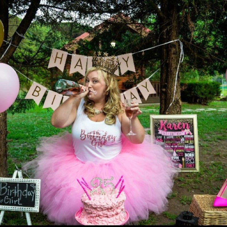 Puede incluir: Una mujer con una falda de tul rosa y una camiseta blanca con el texto "The Birthday Girl" sostiene una copa de vino y una botella de champ&aacute;n. Est&aacute; de pie frente a un pastel de cumplea&ntilde;os rosa y dorado con el n&uacute;mero 35. Detr&aacute;s de ella hay un cartel que dice "Karen" con una lista de cosas que le gustan.
