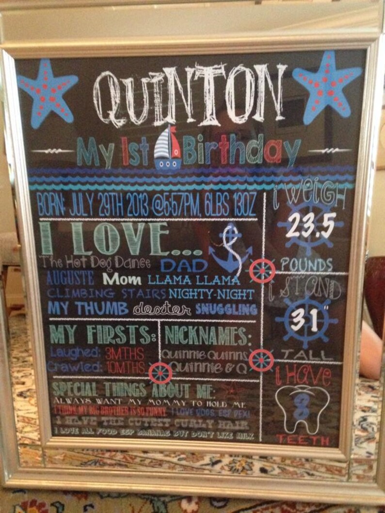 May include: A framed chalkboard sign with the name "Quinton" and "My 1st Birthday" in white chalk. The sign includes details about the child's birth, weight (6 lbs 13 oz), and height (78.7 cm). Decorative elements include blue starfish and a sailboat.