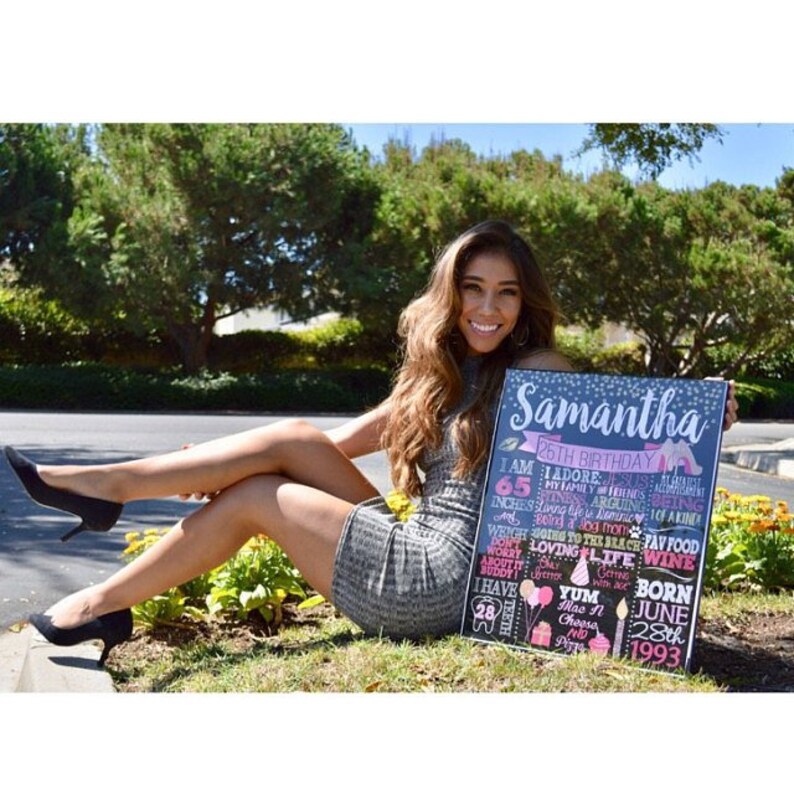 Puede incluir: Un letrero de pizarra con un dise&ntilde;o floral rosa y azul y el texto "Samantha 26th Birthday". El letrero enumera datos sobre Samantha, incluyendo su altura, sus comidas favoritas y su fecha de nacimiento. Una mujer sostiene el letrero y sonr&iacute;e a la c&aacute;mara.
