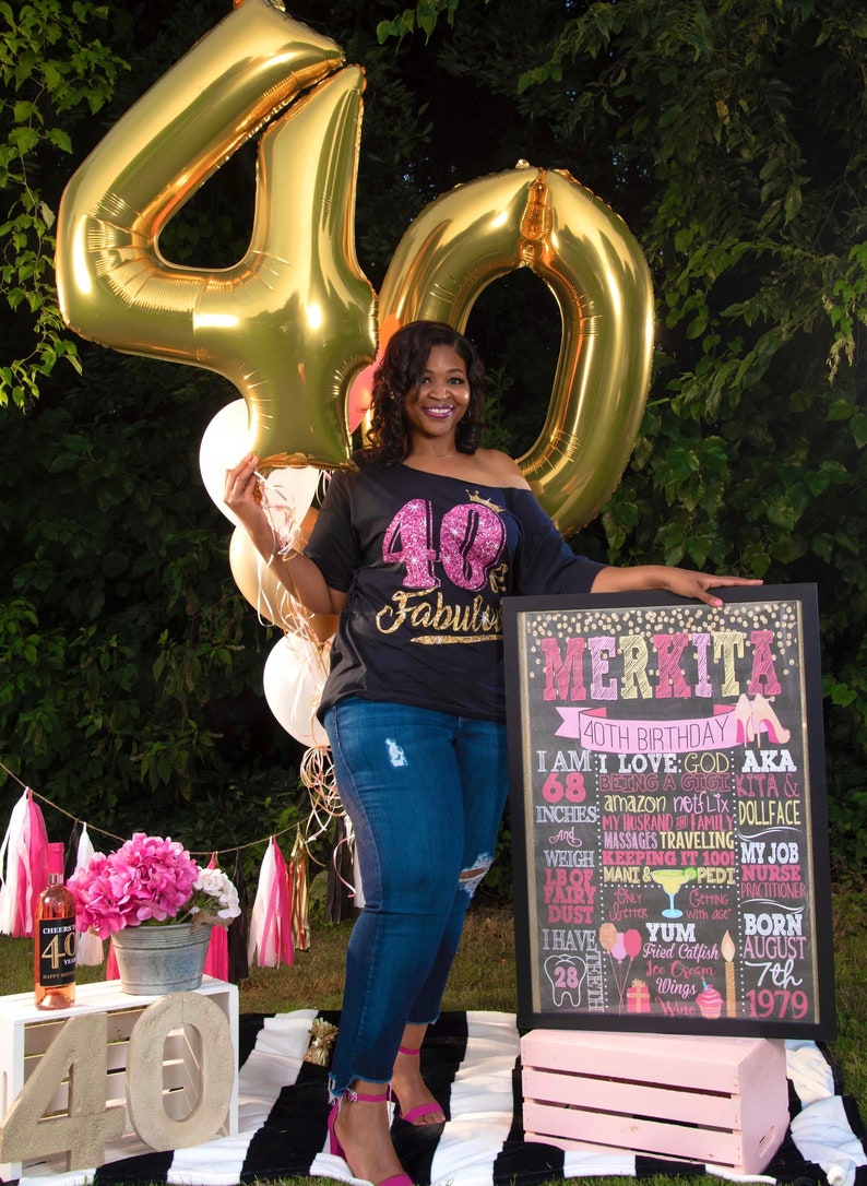 Puede incluir: Una mujer se encuentra de pie frente a un letrero de pizarra negra con detalles de purpurina dorada. El letrero dice "Merkita 40th Birthday" y enumera detalles sobre su vida. Ella sostiene un gran globo dorado en forma del n&uacute;mero 40. Lleva una camiseta negra con purpurina rosa y las palabras "40 & Fabulous".