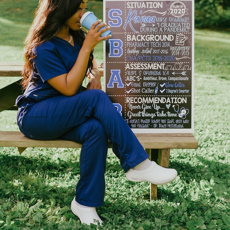 May include: A woman in navy blue scrubs sits on a wooden bench, holding a blue coffee cup. A sign behind her lists "Situation," "Background," "Assessment," and "Recommendation." The sign includes text about graduating during a pandemic.