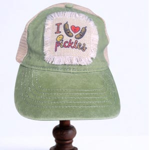 May include: Green trucker hat with a tan mesh back. The front patch reads "I ♥ Pickles" with a heart and pickle graphic. The hat is displayed on a wooden stand.