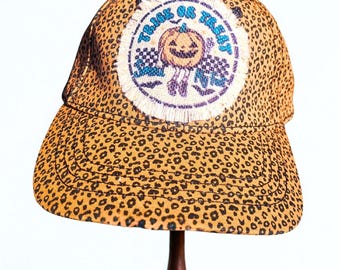 May include: A baseball cap with a leopard print pattern. The cap features a circular patch with a smiling jack-o'-lantern and the words "Trick or Treat". The patch has a frayed edge and is surrounded by blue and white accents.