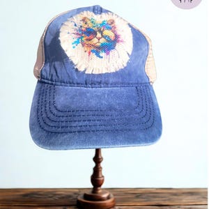 May include: Blue trucker hat with a colorful cat graphic. The hat features a distressed blue front panel, a beige mesh back, and a curved brim. The cat design is a watercolor-style image with a white fringe.