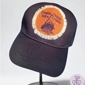 May include: A navy blue baseball cap with a circular patch featuring the text "Double, Double, Toil & Trouble" around a witch's cauldron. The patch has an orange background and a frayed edge.
