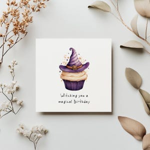 May include: A white card with a purple cupcake topped with a witch's hat. The cupcake has white frosting and gold sprinkles. The text on the card reads "Witching you a magical Birthday."