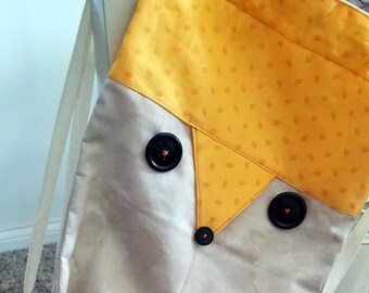 Pode incluir: Uma bolsa de tecido bege e amarela em forma de raposa com uma alça longa. A bolsa tem três botões pretos para os olhos e o nariz.