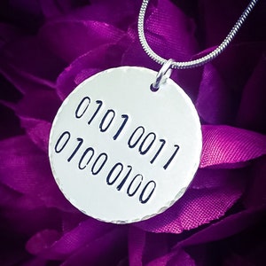 Puede incluir: Un collar de plata con un colgante redondo que presenta el código binario "01010011 01000100" grabado en él. El collar está fotografiado sobre un fondo morado.