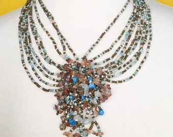 Collar de cuentas de semillas multihilo en oro, azul y beige con medallón de cuentas de cristal.