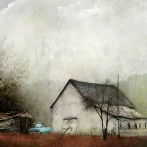 May include: A weathered white barn with a blue truck parked in front of it. The barn has a large, open door and a wooden fence in front. The scene is set in a rural area with trees and a cloudy sky.
