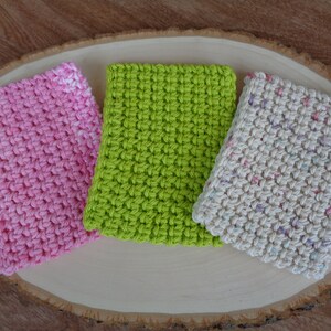 May include: Three crocheted dishcloths in pink, green, and cream with a speckled pattern. The dishcloths are displayed on a wooden surface.