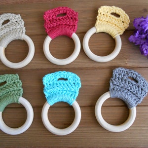 May include: Six wooden teething rings with crocheted tops in various colors, including beige, red, yellow, green, blue, and gray. The rings are arranged in two rows of three on a wooden surface.