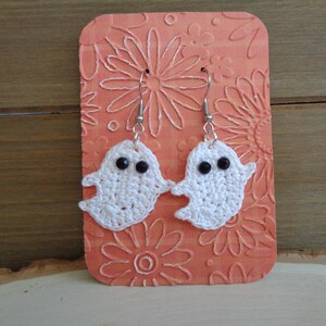 May include: A pair of crocheted ghost earrings with black bead eyes. The earrings are hanging from silver hooks and are displayed on a peach-colored background with a floral pattern.