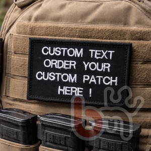 Può includere: Una toppa rettangolare nera con testo ricamato in bianco che recita CUSTOM TEXT ORDER YOUR CUSTOM PATCH HERE !. La toppa è fissata su un gilet tattico color marrone chiaro con cinghie orizzontali e tre tasche nere per caricatori sotto.