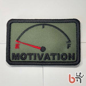Może przedstawiać: Zielona łatka z tkaniny z czarną obwódką. Łatka przedstawia wskaźnik paliwa z czerwoną wskazówką wskazującą na "E" i słowem "MOTIVATION" pod wskaźnikiem.