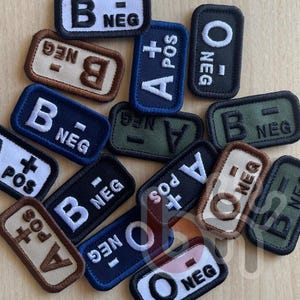 Peut inclure: Une collection d'écussons rectangulaires brodés indiquant divers groupes sanguins, notamment A, B et O, avec des signes positifs et négatifs. Les écussons sont de couleurs variées comme le noir, le bleu marine, le vert olive et le beige, avec des lettres et des bordures brodées en blanc ou en noir.