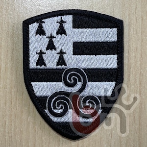 以下が含まれることがあります： 盾形の刺繍ワッペンで、黒と白のデザインが施されています。左上には白い背景に5つの黒いオコジョの斑点があり、右側には黒と白の水平なストライプがあります。下部中央には黒いトリスケリオンのシンボルが配置されています。