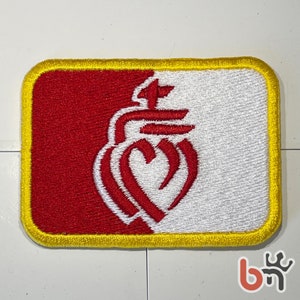 Puede incluir: Un parche bordado rojo y blanco con un diseño de corazón estilizado. El parche tiene un borde amarillo y está hecho de tela.