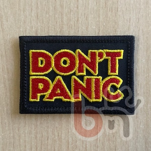 Peut inclure: Un écusson rectangulaire brodé sur fond noir avec une bordure noire épaisse. Le texte DON'T PANIC est cousu en lettres rouges audacieuses avec un contour jaune vif, centré sur l'écusson dans une police de caractères sans empattement.
