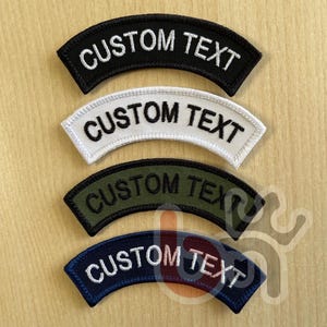 Op de afbeelding: Vier geborduurde patches in zwart, wit, olijfgroen en marineblauw. Elke patch is gebogen en heeft de tekst "CUSTOM TEXT" in een contrasterende kleur. De patches worden gepresenteerd op een licht houten oppervlak.