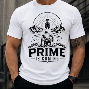 Puede incluir: Una camiseta blanca de manga corta con un estampado gráfico negro que muestra a una persona en cuclillas, un casco espartano, montañas y cadenas rotas. El texto "PRIME IS COMING" aparece impreso en letras mayúsculas negras bajo el gráfico.