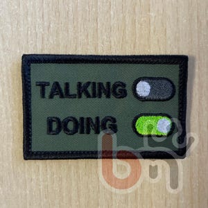 Puede incluir: Un parche rectangular bordado en color verde oliva con borde negro. El texto TALKING está en negro con un interruptor gris en posición de apagado. El texto DOING está en negro con un interruptor verde brillante en posición de encendido.