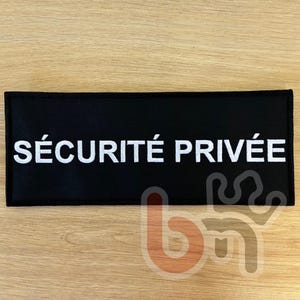 May include: A black rectangular patch with the white text "SÉCURITÉ PRIVÉE". The patch, likely for security, is displayed on a light brown wooden surface. The text is in French.