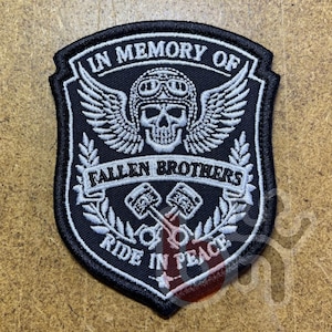 Peut inclure: Écusson brodé noir et blanc en forme de bouclier. L'écusson représente un crâne portant un casque avec des ailes, le texte "IN MEMORY OF" et "FALLEN BROTHERS", et les mots "RIDE IN PEACE".