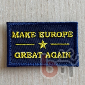 Op de afbeelding: Een rechthoekige donkerblauwe geborduurde patch met de gele tekst "MAKE EUROPE" op de bovenste regel en "GREAT AGAIN" op de onderste regel, gescheiden door een centrale gele ster en twee horizontale lijnen in een vetgedrukt lettertype.