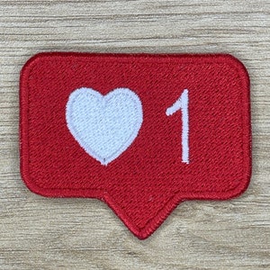 Puede incluir: Parche rojo bordado con un corazón blanco y el número 1. El parche tiene forma de burbuja de diálogo.