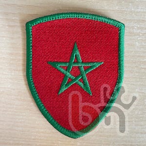 May include: A green embroidered patch shaped like a shield with a red background and a green five-pointed star in the centre.