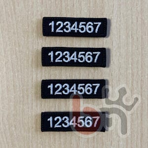 Puede incluir: Cuatro parches rectangulares negros bordados dispuestos verticalmente. Cada parche presenta los números 1234567 bordados en blanco con una fuente sans-serif clara. El parche inferior muestra un ligero tono rosado en los números.