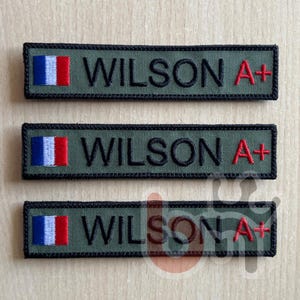 Peut inclure: Trois écussons rectangulaires brodés vert olive avec des bordures noires. Chaque écusson présente un petit drapeau français à gauche, le nom WILSON en lettres capitales noires au centre et le texte rouge A+ sur le côté droit.