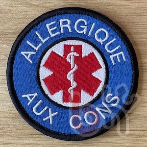 Puede incluir: Un parche redondo con fondo azul y borde negro. El texto "ALLERGIQUE AUX CONS" está en blanco, arqueado en la parte superior e inferior. Un símbolo médico rojo y blanco está en el centro, con un caduceo blanco.