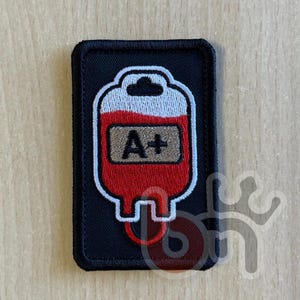 May include: A rectangular embroidered patch with a black background featuring a white and red blood bag graphic. The center of the blood bag contains a tan rectangular label with the black embroidered text A+ centered inside the design.