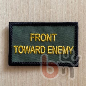 Puede incluir: Un parche rectangular bordado con un fondo verde oliva y un borde negro grueso. El texto FRONT TOWARD ENEMY está cosido en letras mayúsculas amarillas en negrita en el centro del parche, utilizando un estilo de fuente de bloque estándar.