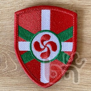 Peut inclure: Écusson brodé en forme de bouclier avec le drapeau basque. Le patch a un fond rouge, une croix blanche et des lignes diagonales vertes. Un cercle blanc au centre contient un symbole lauburu rouge. Pour vêtements ou accessoires.