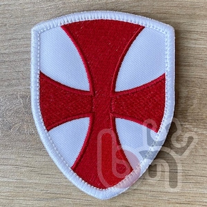 Peut inclure: Écusson en forme de bouclier avec une croix rouge sur fond blanc, bordé d'une couture blanche. Le patch a un design classique, adapté à diverses applications. Le patch est probablement en tissu.