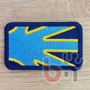 Puede incluir: Parche rectangular con fondo azul y diseño geométrico amarillo y azul. El parche tiene un borde azul oscuro. El diseño presenta líneas en zigzag amarillas y un fondo azul.