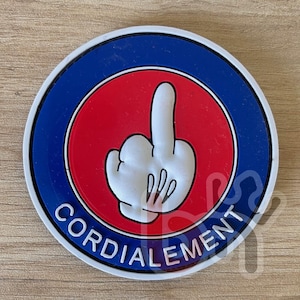 May include: A round coaster with a blue and white border, a red center, and a white hand giving the middle finger. The word "CORDIALEMENT" is printed in white around the bottom of the coaster.