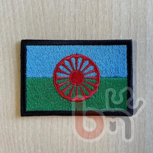 May include: Embroidered patch featuring the Romani flag. The patch has a rectangular shape with a black border. The top half is light blue, the bottom half is green, and a red wheel is in the center.
