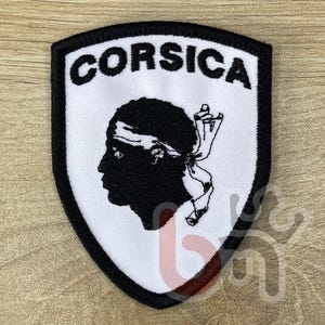 Puede incluir: Parche bordado con la palabra "CORSICA" sobre una silueta negra de una cabeza con una bandana blanca. El parche tiene un fondo blanco y un borde negro, con forma de escudo.