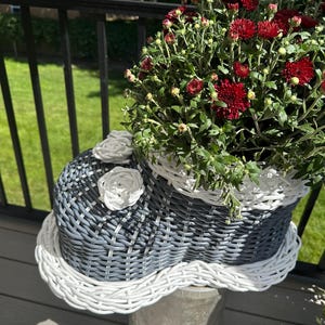 May include: A shoe-shaped wicker planter featuring a dark gray woven body with a white woven rim and base. Two white woven flower accents are attached to the top of the shoe. The planter is filled with a dense cluster of deep red chrysanthemum flowers.