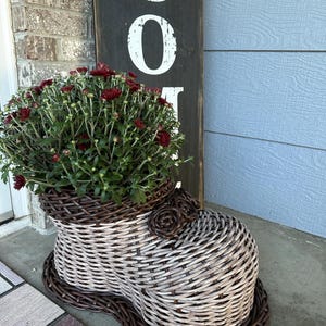 May include: A woven wicker planter shaped like a boot with a two-tone light gray and dark brown finish. The planter features decorative woven rosettes on the top and is filled with a dense cluster of dark red chrysanthemum flowers.