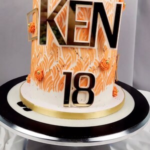 May include: A birthday cake with orange and white frosting, decorated with the name "KEN" and the number "18" in gold and black. A decorative controller sits atop the cake.