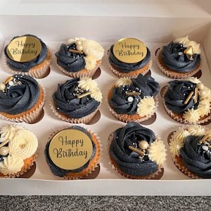 May include: A box of cupcakes decorated with black and white frosting and gold sprinkles. The cupcakes have gold round toppers with the text "Happy Birthday" on them.