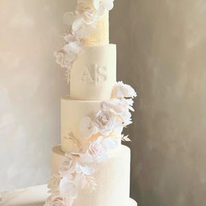 May include: A four-tiered white wedding cake decorated with cascading white flowers. The cake has a smooth finish with the initials "AS" embossed on the second tier. The flowers include roses and orchids.