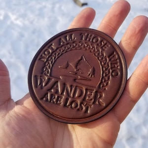 Puede incluir: Un posavasos redondo de cuero marrón con la frase en relieve "NOT ALL THOSE WHO WANDER ARE LOST" que rodea un diseño de montaña y árbol. El posavasos se sostiene en una mano sobre un fondo nevado.