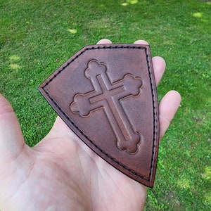 May include: A brown leather shield with a cross embossed in the center. The shield has a stitched edge.